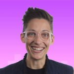 Headshot of a queer person with olive skin. Big eyes. Bigger grin. Pink frames. Pompadour. Quilted blue vest armour.