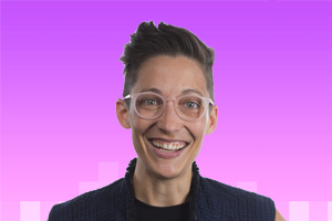 Headshot of a queer person with olive skin. Big eyes. Bigger grin. Pink frames. Pompadour. Quilted blue vest armour.