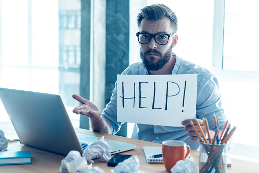 hipster at laptop looking flustered and holding up a help sign, meant to illustrate he needs help with accessibility compliance